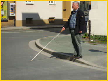 Der blinde Verkehrsteilnehmer signalisiert durch vorstrecken des Blindenlangstockes auf die Fahrbahn, dass er nun die Straße überqueren wird.